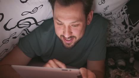 Close-up-of-smiling-face-of-a-young-bearded-and-mustashed-man,-using-digital-tablet-under-blanket,-in-his-bed,-at-night.-His-yorkshire-terrier-is-lying-next-to-him