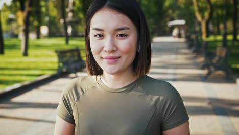 Retrato-al-aire-libre-de-la-joven-asiática,-escuchando-música-en-los-airpods-y-sonriendo-a-la-cámara-en-el-parque,-el-seguimiento-de-la-toma