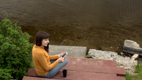 Una-joven-disfruta-de-un-teléfono-sentado-en-un-muelle-junto-al-estanque-en-el-verano.