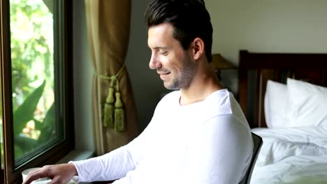 Young-Man-Using-Laptop-Computer-Happy-Smiling-Guy-Chatting-Online-Over-Big-Window-With-Tropical-Garden-View