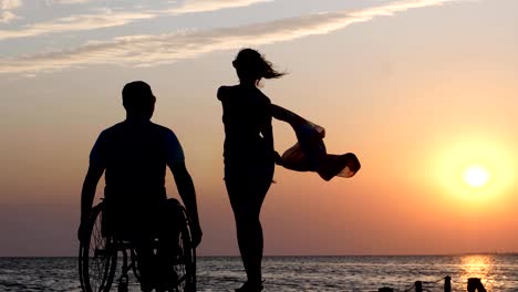 Woman-is-standing-on-jetty-and-waving-a-cloth-in-front-of--a-disabled-man-in-a-wheelchair,-background-of-orange-sundown-over-river