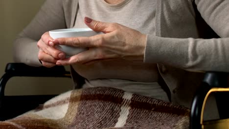 Mature-woman-sitting-near-window-in-wheelchair-drinking-hot-tea,-home-coziness