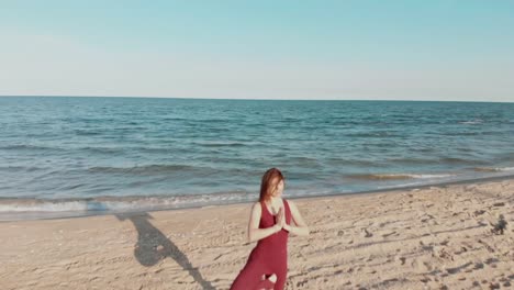 Drone-bewegt-sich-hinter-Frau-in-Sport-Wear-hält-Yoga-Asana-Position-am-Meer-oder-Ozean-Sandstrand.-Windige-sonniges-Wetter.-Luftaufnahme-des-friedlichen-Gesundheit-Mädchen-Praxis-durchführen