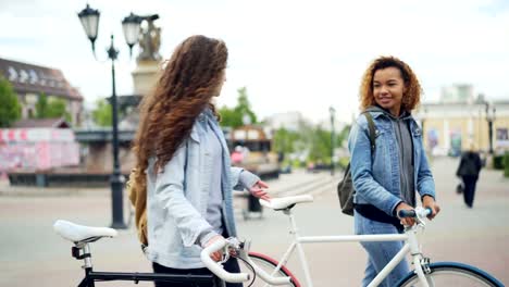 Cheerful-young-women-are-talking-while-walking-with-bikes-along-beautiful-street-with-fountain-and-beautiful-buildings-and-people-moving-in-background.