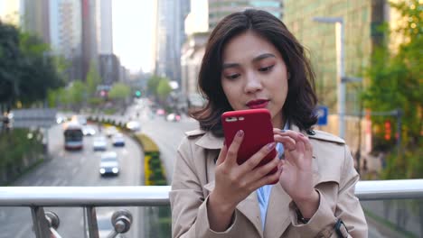 Bastante-feliz-joven-mujer-asiática-mediante-teléfono-móvil-en-la-ciudad-China-de-Chengdu,-en-la-tarde-en-el-puente-con-la-carretera-muy-transitada-en-el-fondo