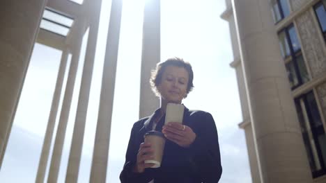 Tracking-left-shot-of-middle-adult-female-manager-with-short-hair-standing-outdoors-on-windy-day,-holding-coffee-cup-and-texting-on-her-mobile-phone-with-smile