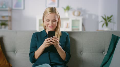 Hermosa-rubia-mujer-utiliza-teléfono-móvil-mientras-está-sentado-en-un-sofá-en-casa.-Sonriendo-feliz-mujer-utiliza-el-Smartphone-para-navegar-a-través-de-Internet,-redes-sociales-y-viendo-Videos.