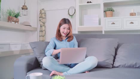 Portrait-of-beautiful-attractive-young-smiling-Asian-woman-using-computer-or-laptop-while-lying-on-the-sofa-when-relax-in-living-room-at-home.-Enjoying-time-lifestyle-women-at-home-concept.