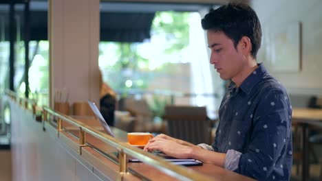 Attractive-hispanic-good-looking-young-Latin-American-man-sitting-in-coffee-shop-working,-writing,-typing-on-laptop-and-drinking-coffee,-medium-shot