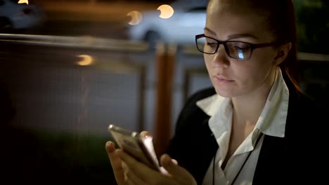 Retrato-de-la-joven-mujer-caucásica-hermosa-usando-la-mano-del-teléfono-inteligente-mantener-al-aire-libre-en-la-noche-de-la-ciudad,-sonriendo,-cara-iluminada-ScreenLight-red-social,-tecnología,-concepto-de-comunicación