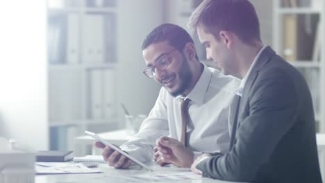 Two-Cheerful-Businessmen-Using-Tablet-and-Having-Discussion-in-Office