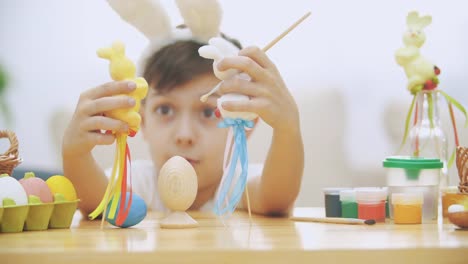 Young-adorable-boy-is-sitting-at-the-table-full-of-Easter-decorations-and-is-playing-with-Easter-bunnies-in-his-hands.-Bunnies'-discussion.-Who-would-paint-an-egg-Bunny-theatre.