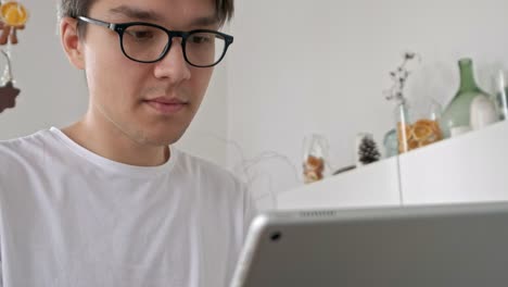 Attractive-man-at-home-using-tablet-in-kitchen-sending-message-on-social-media-smiling-enjoying-modern-lifestyle-wearing-white-shirt