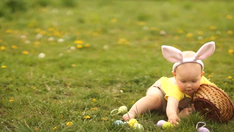 Schönes-Baby-in-einem-Osterhasen-Kostüm-sammelt-Ostereier-in-einem-Korb,-der-auf-dem-Rasen-im-Park-sitzt.-Frühlingshaftes,-glückliches-Osterfest