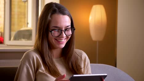 Closeup-portrait-of-young-attractive-caucasian-female-in-glasses-using-the-tablet-smiling-happily-sitting-on-the-couch-indoors-in-apartment