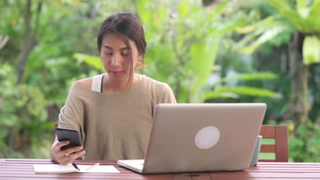 Freelance-Asian-woman-working-at-home,-business-female-working-on-laptop-and-using-mobile-phone-drinking-coffee-sitting-on-table-in-the-garden-in-morning.-Lifestyle-women-working-at-home-concept.