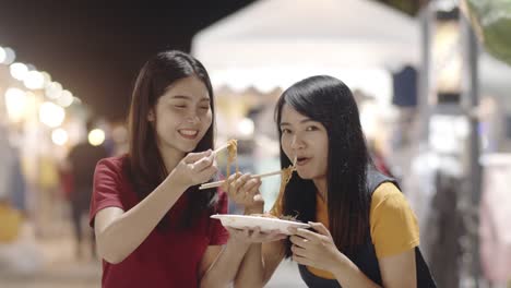 Asian-young-women-friends-travel-in-Bangkok,-Thailand,-beautiful-female-feeling-happy-walking-and-eating-Pad-Thai-at-The-Khao-San-Road.-Women-travel-eat-street-food-concept.-Slow-motion-shot.