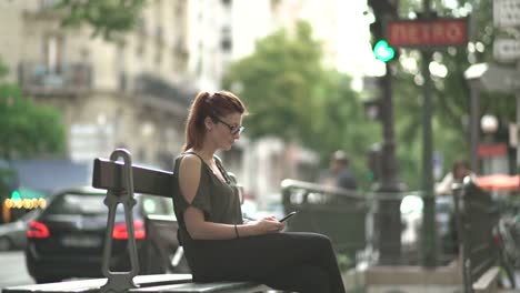 Attraktive-kaukasisch-nachdenkliche-Frau-mit-Brille,-Sommersprossen,-Piercings-und-roten-Haaren-warten-und-beobachten-an-ihrem-Smartphone-sitzen-auf-der-Bank,-während-sonnigen-Sommer-in-Paris.-4K-UHD.