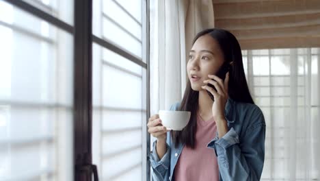 Atractivo-retrato-sonriente-joven-asiática-mujer-bebe-café-y-hablando-por-teléfono-con-amigos-de-pie-al-lado-de-la-ventana-en-la-oficina-en-casa.