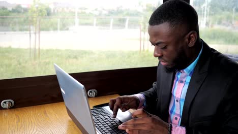 Black-businessman-is-working-typing-a-message-on-laptop-sitting-in-summer-cafe.