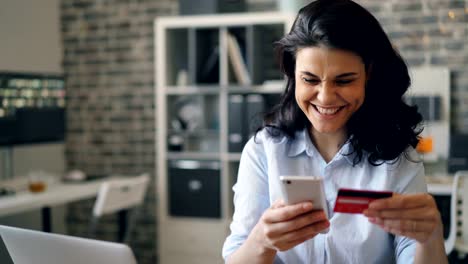 Joyful-girl-shopping-online-paying-with-credit-card-using-smartphone-in-office