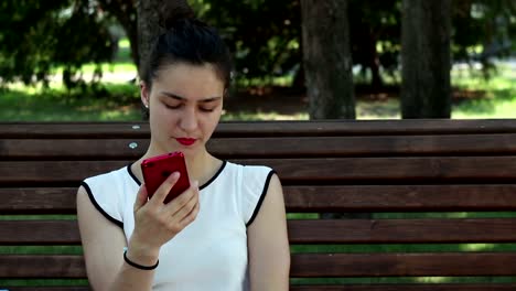 Retrato-de-cerca-Una-hermosa-joven-con-una-camiseta-blanca-está-chateando-en-las-redes-sociales-en-su-teléfono-inteligente-mientras-está-sentada-en-un-banco-en-un-parque.