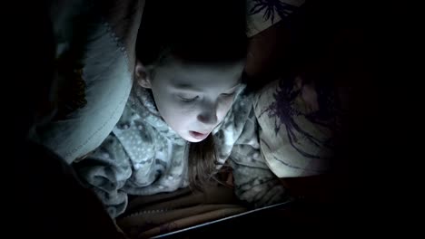 Cute-little-girl-at-night-in-the-dark-playing-on-the-tablet-under-the-blanket-on-the-bed.-Concept-video.-Close-up.-Raw-video.-4K.