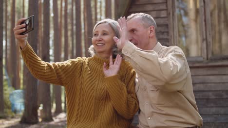 Pareja-vieja-teniendo-videollamada-cerca-de-la-casa-de-madera