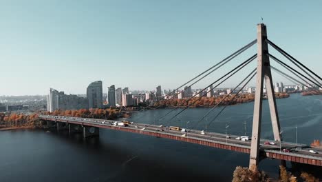 Betonbrücke-mit-Autos-über-den-Fluss-in-der-Metropole.-Drohne-fliegt-in-der-Nähe-der-Großstadtbrücke-mit-Hochhäusern-im-Hintergrund.-Luftaufnahme-des-Stadtzentrums-mit-Fluss-und-Brücke.-Kiew,-Ukraine