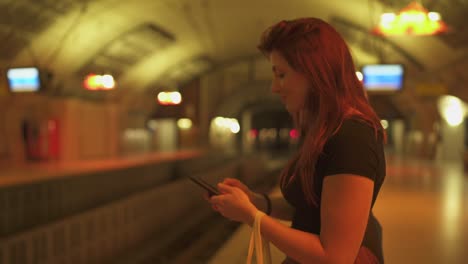 Atractiva-mujer-pelirroja-con-pecas,-piercings-y-pelo-rojo-charlando-en-el-teléfono-inteligente-en-la-estación-de-metro,-durante-el-verano-soleado-en-París.-Fondo-subterráneo-borroso.-4K-UHD.