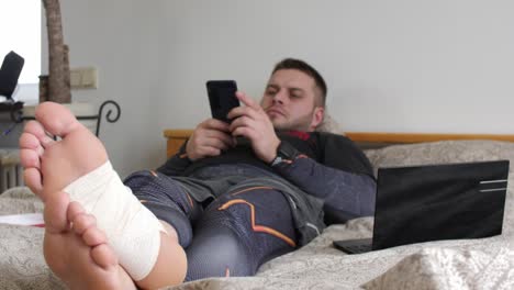 Bearded-man-with-injured-leg-using-his-smartphone-in-the-bedroom