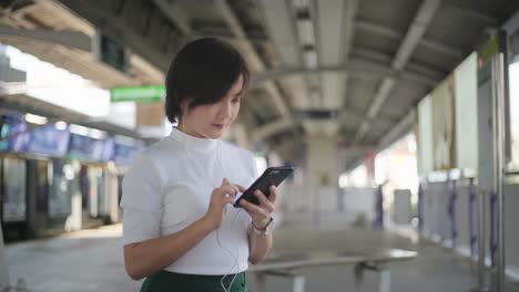 Retrato-de-mujer-asiática-con-auriculares-escuchando-música-y-usando-teléfono-inteligente-para-chatear-con-amigos-o-navegar-mientras-espera-un-tren.-Tecnología-en-la-vida-cotidiana-y-los-viajes.