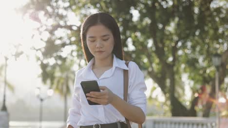 Schöne-asiatische-Geschäftsfrau-in-einem-weißen-Hemd-verwendet-ein-Smartphone-SMS-Teilen-von-Nachrichten-auf-Social-Media,-während-außerhalb-des-Morgens-in-einem-öffentlichen-Park.