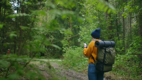 Cámara-lenta:-Joven-excursionista-usando-su-teléfono-inteligente-mientras-disfruta-de-su-fin-de-semana-en-el-bosque.-Mujer-viajera-con-mochila-caminando-por-el-camino-del-bosque-mirando-el-teléfono-y-mapa-en-madera-verde