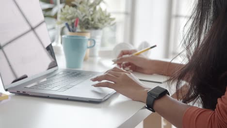 Close-up-hands-asian-businesswoman-using-a-laptop-and-write-on-a-book-at-the-home-office.