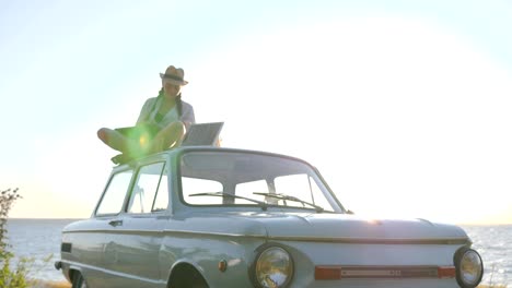 natural-energy,-girl-sits-on-roof-blue-classic-car-with-solar-battery-and-laptop-in-hands-in-open-air,-female-sitting-on-car-rooftop