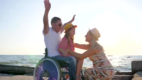 little-girl-sitting-on-hands-paralyzed-daddy-in-wheelchair-with-outstretched-arms-in-backlight-pregnant-wife-with-child-and-disabled