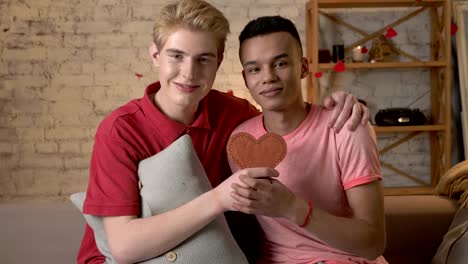 Multiethnic-happy-gay-couple-sitting-on-the-couch-and-holding-a-heart-sign-in-hands.-Homeliness,-LGBT-loverHappy-s,-happy-gay-family-concept.-60-fps