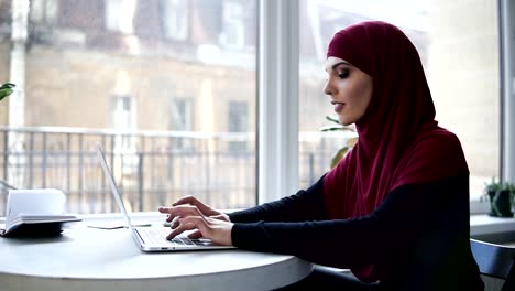 Young-attractive-muslim-girl-with-hijab-covering-her-head-is-typing-something-on-her-laptop-while-sitting-in-some-supposedly-co-working-space-with-glass-windows-on-the-background