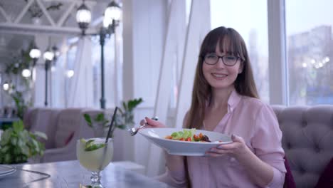 gesunder-Lebensstil,-Mädchen-mit-langen-Haaren-in-Brillen-mit-einer-Gabel-und-Teller-in-der-hand-griechischen-Salat-essen-und-Blick-in-die-Kamera