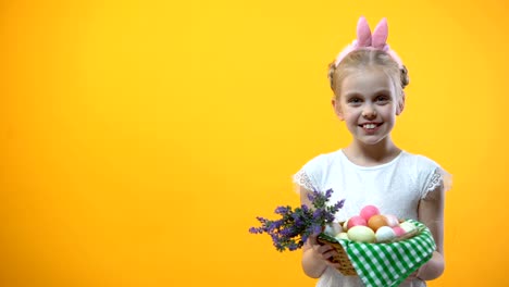 Lächelnde-Kind-zeigt-Körbchen-mit-bunten-Eiern-gelben-Hintergrund,-Ostern