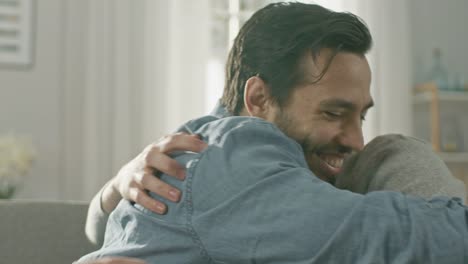 Pareja-Gay-masculino-atractivo-lindo-sentarse-juntos-en-un-sofá-en-casa.-Novios-son-Hugging-y-abrazándose-unos-a-otros.-Ellos-son-feliz-y-sonriente.-Son-vestidos-casualmente-y-sala-tiene-un-Interior-moderno.