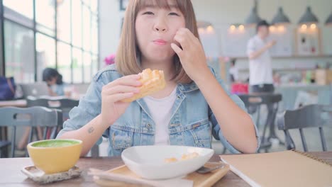 Business-freelance-Asian-woman-eating-bread-and-drinking-warm-cup-of-coffee-while-sitting-on-table-in-cafe.-Lifestyle-smart-beautiful-female-relax-in-coffee-shop-concepts.