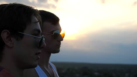 Profile-of-handsome-gay-boys-in-sunglasses-standing-on-the-edge-of-rooftop-and-talking.-Young-male-couple-relaxing-on-roof-of-high-rise-building-and-enjoying-beautiful-cityscape.-Close-up-Slow-motion