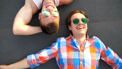 Top-view-of-handsome-gay-boys-in-sunglasses-lying-on-rooftop-of-high-rise-building-and-laughing.-Young-happy-male-couple-looking-at-camera-and-candid-smiling-on-roof.-Crane-shot-Close-up-Slow-motion