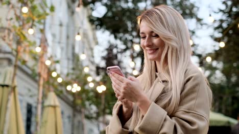 Portrait-of-woman-in-autumn-clothes-writing-message-on-her-smartphone-outdoors.-Girl-using-digital-gadget,-scrolls-through-social-media-on-device,-reading-news-on-app.-Lady-looking-at-camera