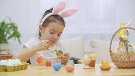 Pequeña-niña-juguetona,-con-orejas-de-conejo-en-la-cabeza-eligió-un-color-naranja-para-pintar-un-huevo.-La-chica-está-masticando-y-saliendo-de-una-mancha-amarilla-en-su-nariz.