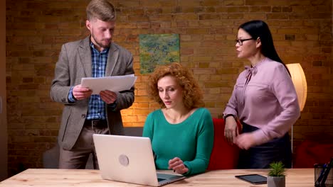 Closeup-Shooting-von-jungen-kaukasischen-Geschäftsfrau,-die-am-Laptop-arbeitet-und-mit-zwei-Kollegen,-die-glücklich-lächeln,-über-Daten-diskutiert.-Mitarbeiterinnen-mit-Tablet