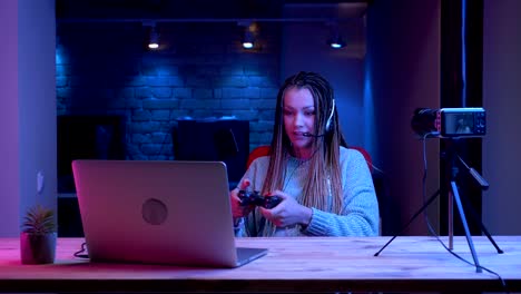 Closeup-shoot-of-young-attractive-female-blogger-with-dreadlocks-in-headphones-playing-video-games-on-the-laptop-with-the-neon-background-indoors