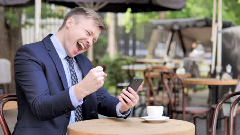 Outdoor-Sitting-Geschäftsmann-feiert-Ergebnisse-auf-Smartphone